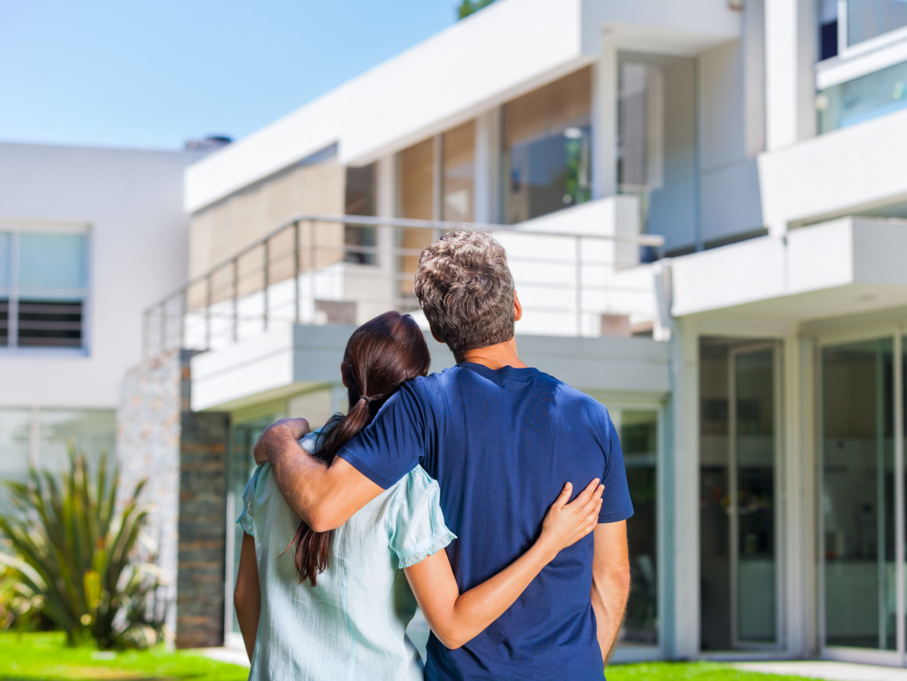 couple embracing in front of new big modern house, outdoor rear view back looking at their dream home